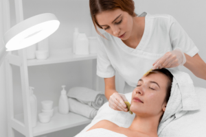 Aud dem Bild sind zwei Frauen zu sehen. Die Kundin liegt auf einer Liege und hat ihre Augen geschlossen. Die Kosmetikerin beginnt die Hautpflege mit zwei grünen Jadesteinen.