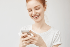 Eine junge rothaarige Frau freut sich über neue Einverständniserklärungen und Pflegehinweise auf unserer Beautymedia Website. Sie hält ein Handy in der Hand.