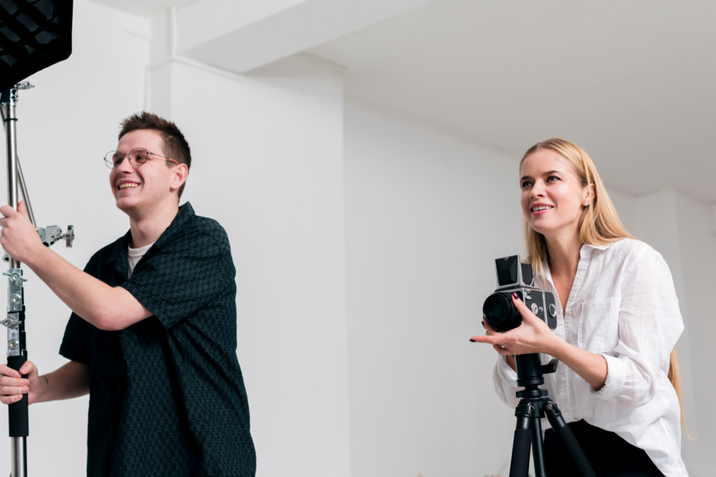 rechts im Bild ist eine blonde junge Dame mit einer Kamera in der Hand. Links im Bild ein Herr der ein Studiolicht hält. Sie könnten neuen Produktbilder für eine Webiste fotografieren.