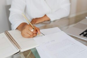 Close-up of a woman's hand writing notes in a planner on a glass desk setup.