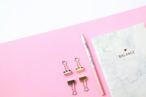 Flat lay of office supplies on a pink background featuring a notebook, pencil, and binder clips.