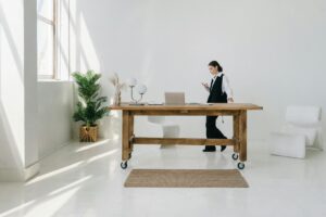 Elegant minimalist office interior with wooden table, potted plant, and woman using smartphone.